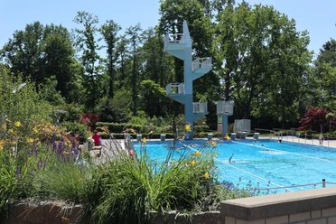 Ein Freibad mit Sprungturm, umgeben von Bäumen und bunten Blumen am Beckenrand.