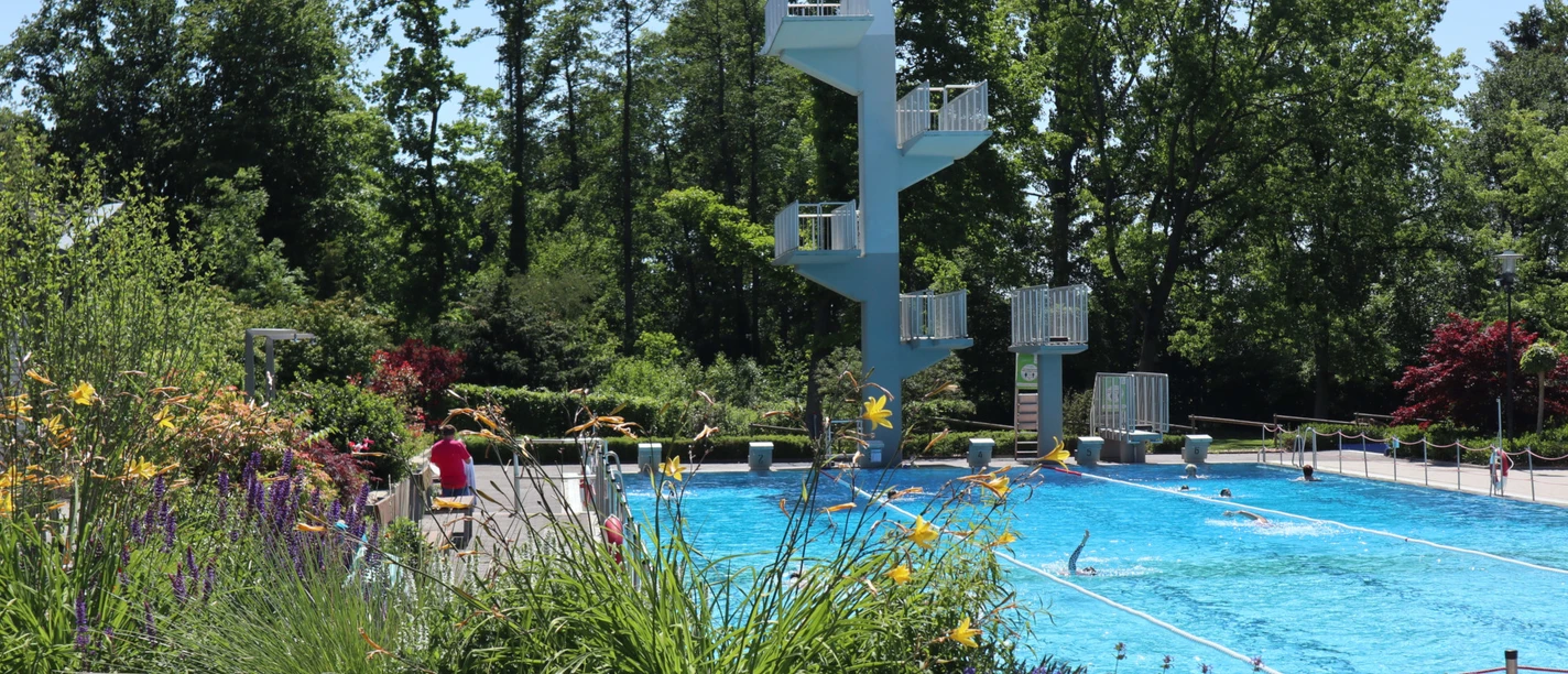 Ein Freibad mit Sprungturm, umgeben von Bäumen und bunten Blumen am Beckenrand.