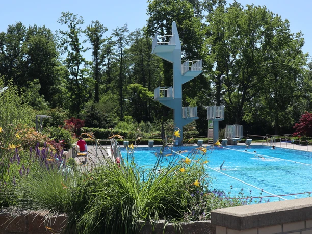 Ein Freibad mit Sprungturm, umgeben von Bäumen und bunten Blumen am Beckenrand.
