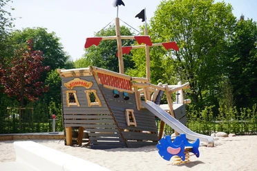 Das neue Piratenschiff im Kinderspielbereich Holzspielplatz in Form eines Piratenschiffs mit Rutsche, Sandboden; umgeben von grünen Bäumen.