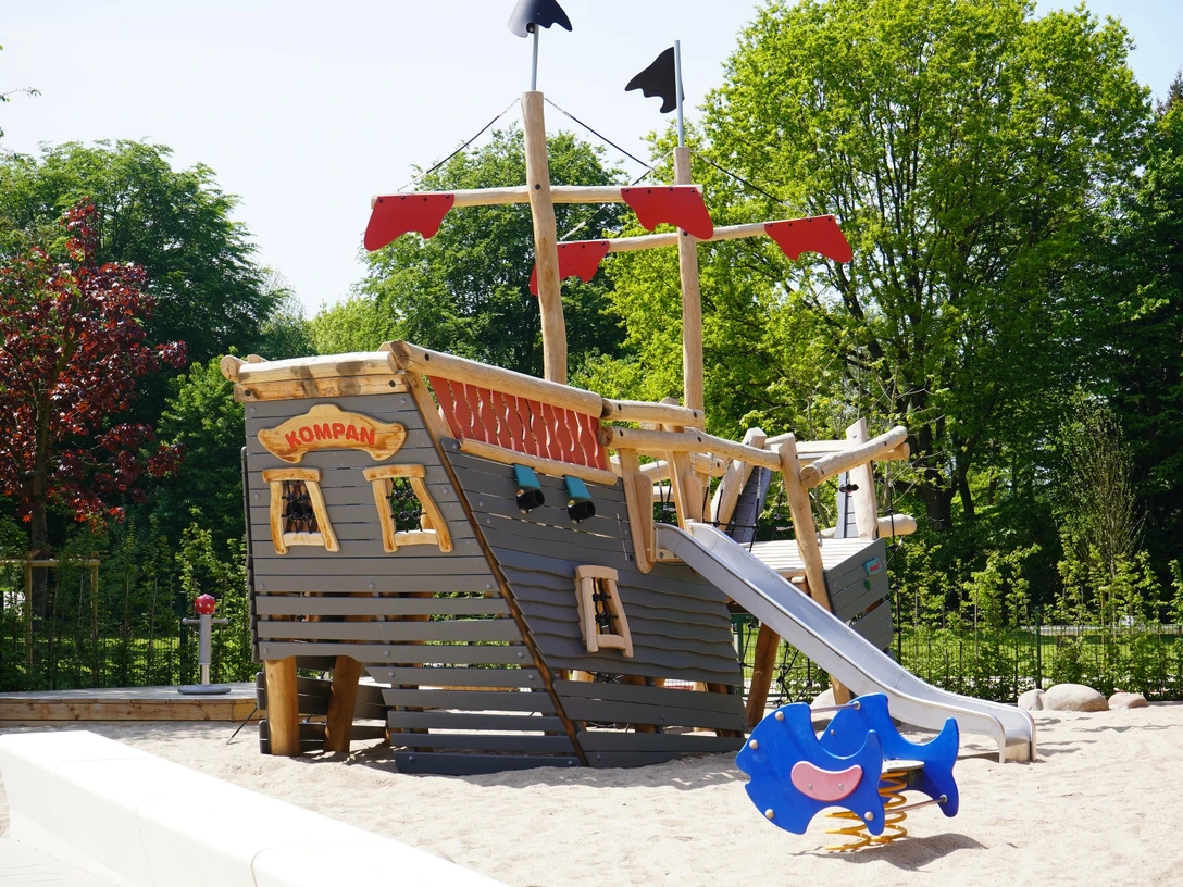Das neue Piratenschiff im Kinderspielbereich Holzspielplatz in Form eines Piratenschiffs mit Rutsche, Sandboden; umgeben von grünen Bäumen.