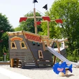 Das neue Piratenschiff im Kinderspielbereich Holzspielplatz in Form eines Piratenschiffs mit Rutsche, Sandboden; umgeben von grünen Bäumen.
