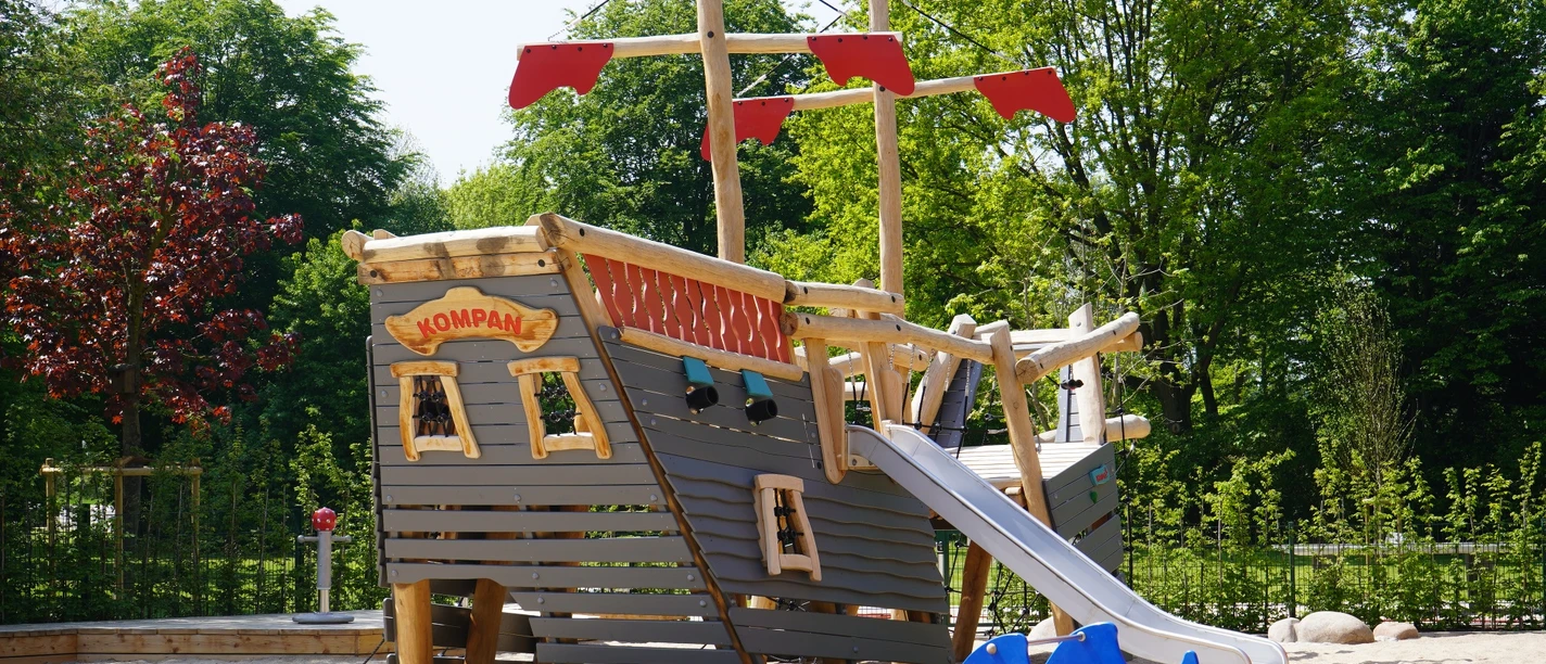 Das neue Piratenschiff im Kinderspielbereich Holzspielplatz in Form eines Piratenschiffs mit Rutsche, Sandboden; umgeben von grünen Bäumen.