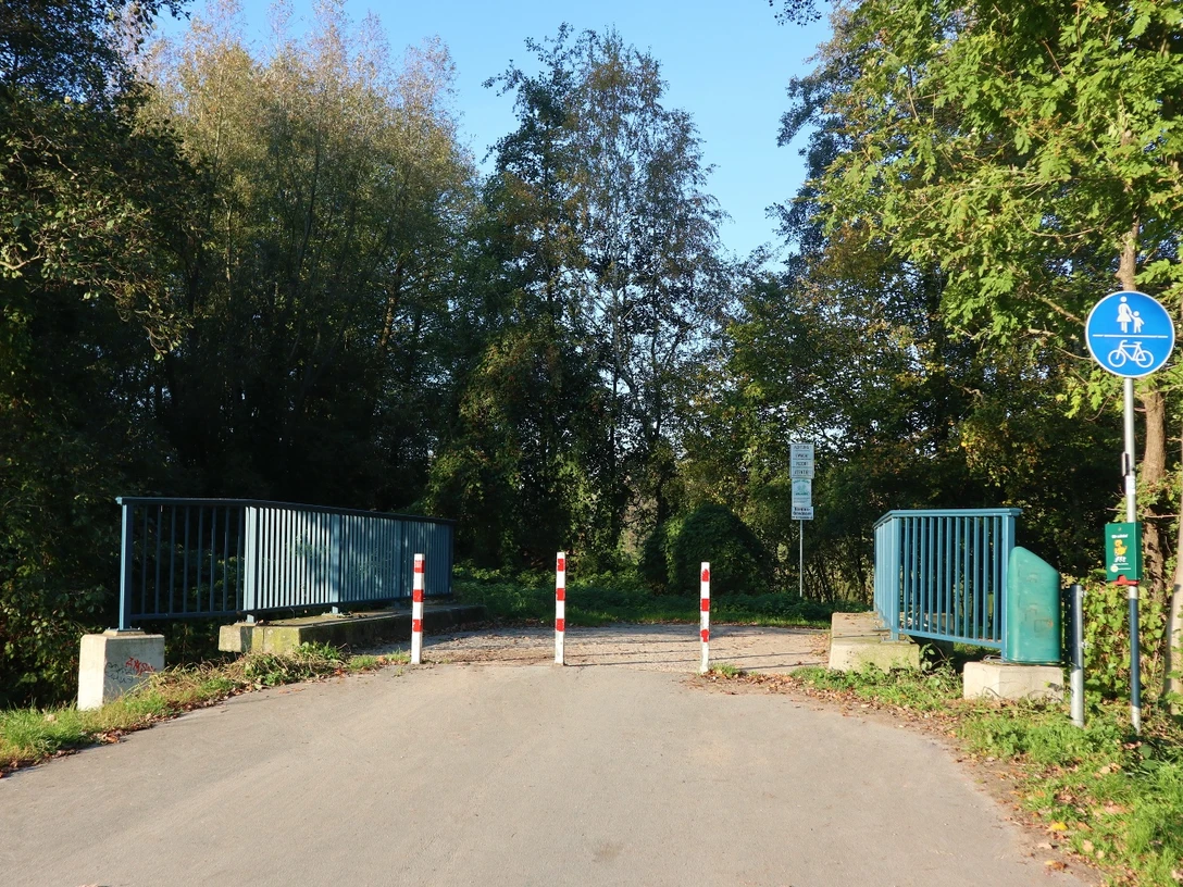 Ein geteilter Rad- und Fußweg führt über eine Brücke, gesäumt von Bäumen, mit Schranken am Eingang.