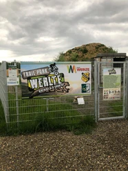 Trail-Park-Werlte Eingang des Trail-Park Werlte mit Infotafeln, Stahlzaun und Hügel im Hintergrund unter grauem Himmel.