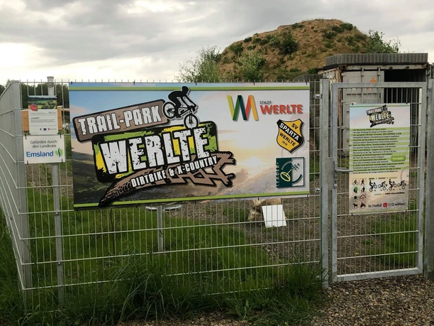 Trail-Park-Werlte Eingang des Trail-Park Werlte mit Infotafeln, Stahlzaun und Hügel im Hintergrund unter grauem Himmel.