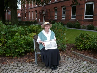 Eine ältere Frau sitzt auf einer Bank, hält eine alte Fotografie vor einem historischen Schulgebäude.
