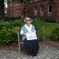 "Bürgerin Gesine" vor dem Gymnasium am Markt (früher Marktschule) Eine ältere Frau sitzt auf einer Bank, hält eine alte Fotografie vor einem historischen Schulgebäude.