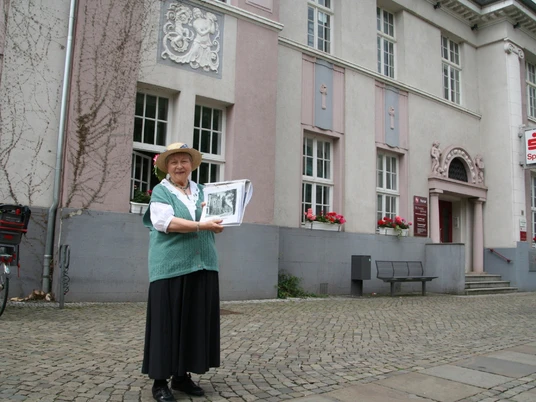 Gesine, eine ältere Frau in traditioneller Kleidung, steht lächelnd vor der Sparkasse Achim.