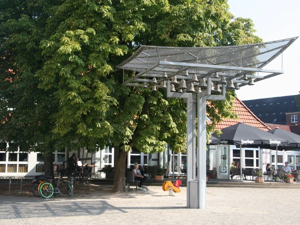 Ein modernes Glockenspiel steht auf einem gepflasterten Platz, umgeben von Bäumen und Menschen.