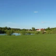 Grüne Landschaft mit Golfplatz im Vordergrund, einem kleinen See und einem roten Gebäude im Hintergrund.