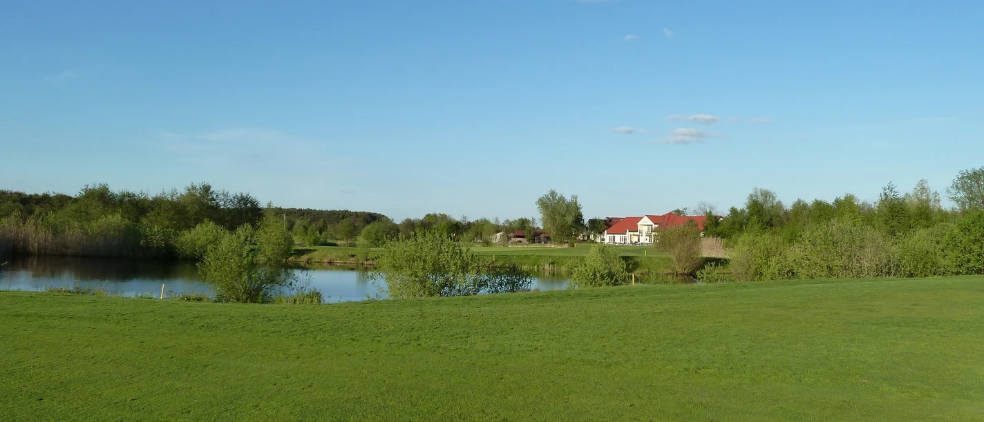 Golfplatz Achim Grüne Landschaft mit Golfplatz im Vordergrund, einem kleinen See und einem roten Gebäude im Hintergrund.