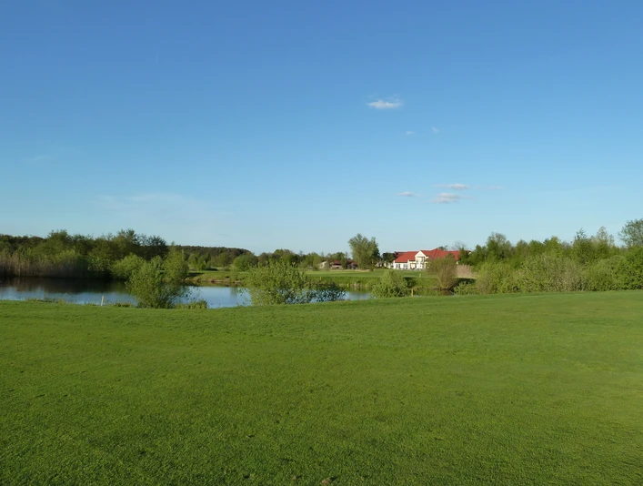 Golfplatz Achim Grüne Landschaft mit Golfplatz im Vordergrund, einem kleinen See und einem roten Gebäude im Hintergrund.