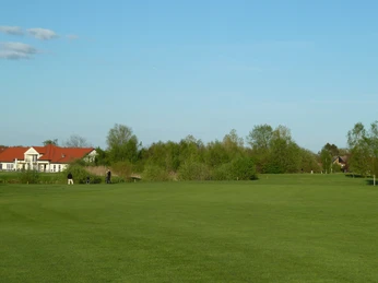 Weitläufiger Golfplatz mit gepflegtem Grün, eingerahmt von Bäumen und einem Gebäude mit rotem Dach.Extensive golf course with manicured greenery, framed by trees and a building with a red roof.Omfattende golfbane med velplejede grønne områder, indrammet af træer og en bygning med rødt tag.Uitgestrekte golfbaan met verzorgd groen, omringd door bomen en een gebouw met een rood dak.