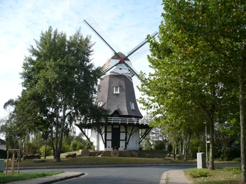 Achimer Windmühle Achimer Windmühle mit Reetdach und imposanten Flügeln umgeben von Bäumen bei klarer Wetterlage.