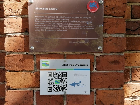 Eine Infotafel aus Metall und ein Schild mit QR-Code sind an einer Backsteinmauer angebracht.