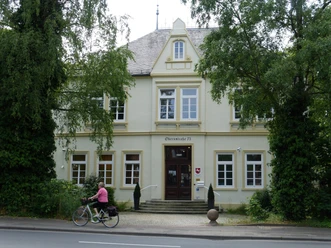 Zweistöckiges Gebäude mit gelber Fassade, üppiges Grün, vorbei radelnde Person auf der Straße.