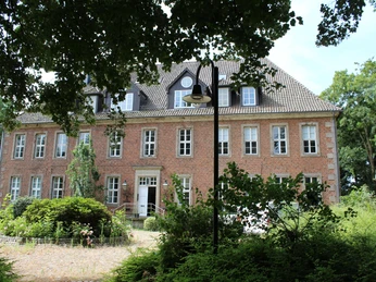 Backsteinfassade eines historischen Gebäudes mit weißen Fensterrahmen und gepflegtem Vorgarten.