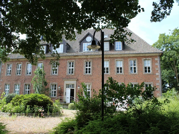 Backsteinfassade eines historischen Gebäudes mit weißen Fensterrahmen und gepflegtem Vorgarten.