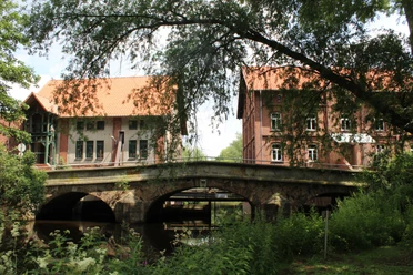 Die Aue-Brücke in Steyerberg, umgeben von Bäumen, verbindet historische Backsteingebäude über einen Fluss.