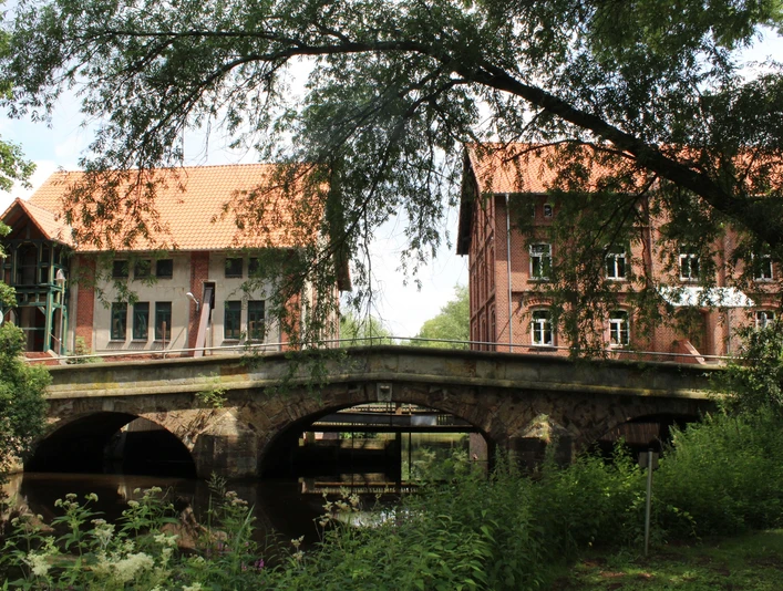 Die Aue-Brücke in Steyerberg, umgeben von Bäumen, verbindet historische Backsteingebäude über einen Fluss.