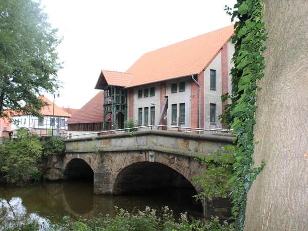 Eine Steinbrücke mit zwei Bögen überspannt einen Fluss, umgeben von Bäumen und historischen Gebäuden.