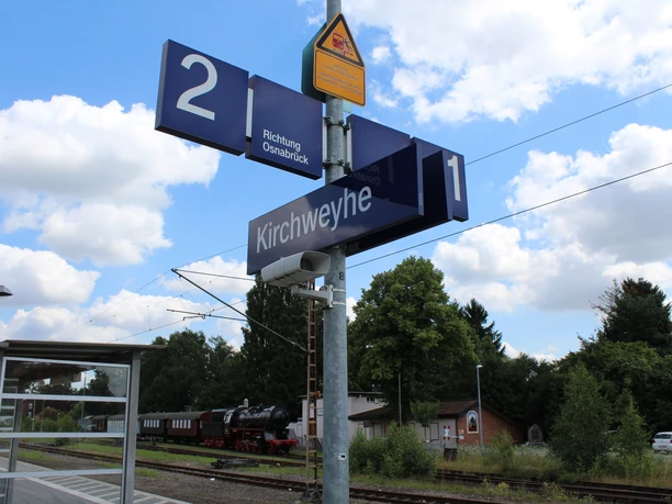 Hinweisschilder am Kirchweyher Bahnhof mit Gleisen und dampfender Lokomotive im Hintergrund.