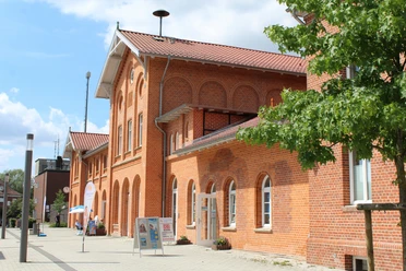 Backsteinbahnhof mit markanten Bögen und roten Dachziegeln in sonniger Umgebung, Bäume im Vordergrund.