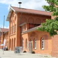 Backsteinbahnhof mit markanten Bögen und roten Dachziegeln in sonniger Umgebung, Bäume im Vordergrund.