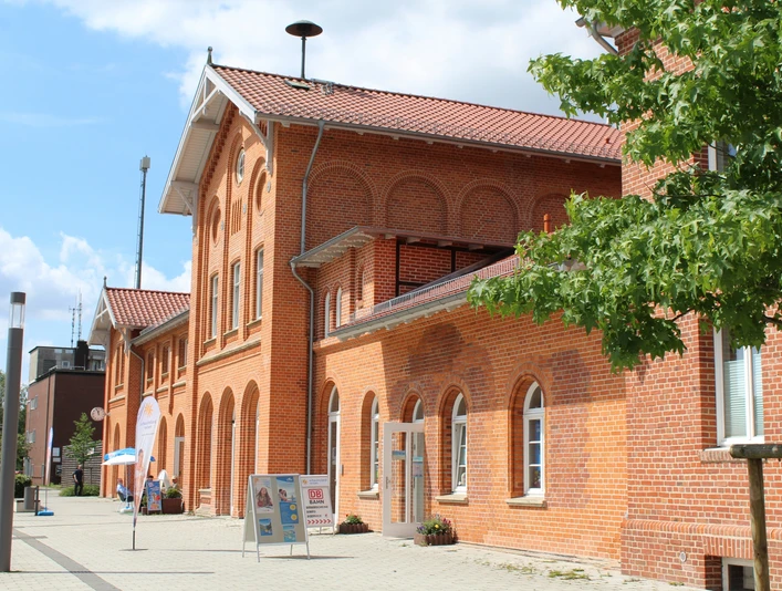 Backsteinbahnhof mit markanten Bögen und roten Dachziegeln in sonniger Umgebung, Bäume im Vordergrund.