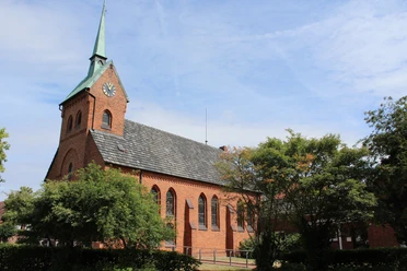 Backsteinkirche mit Spitzdach und Turm im Dorf Bruchhausen; von Bäumen umgeben, blauer Himmel.