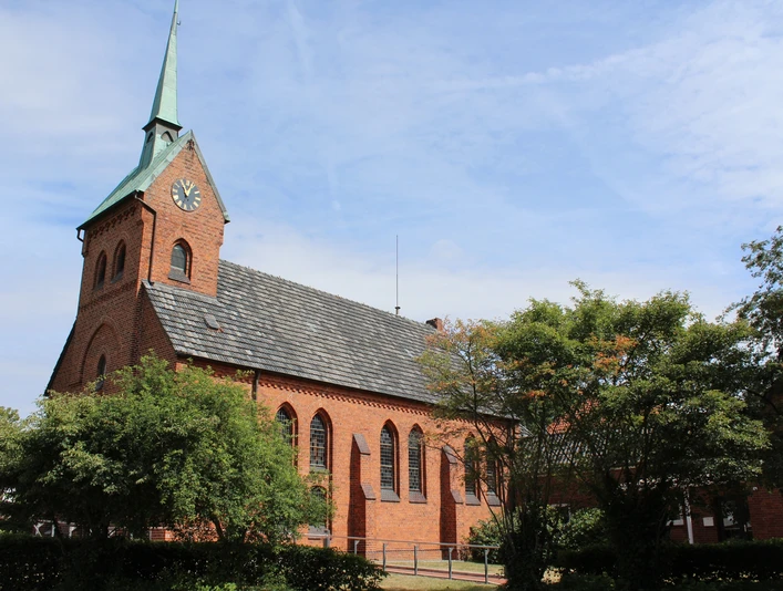 Backsteinkirche mit Spitzdach und Turm im Dorf Bruchhausen; von Bäumen umgeben, blauer Himmel.