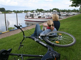In der Marina-Mehlbergen sind Boote am Steg zu sehen. Zwei Menschen entspannen auf einer Wiese, im Vordergrund liegt ein Fahrrad.