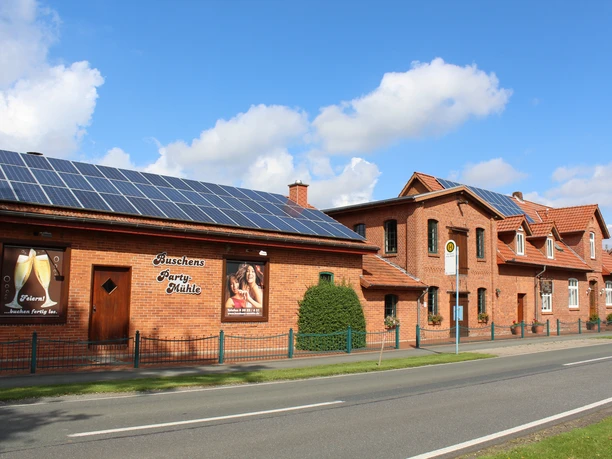Buschens Mühle Nordholz, ein rot verklinkertes Gebäude mit Solarzellen, an einer ruhigen Straße.