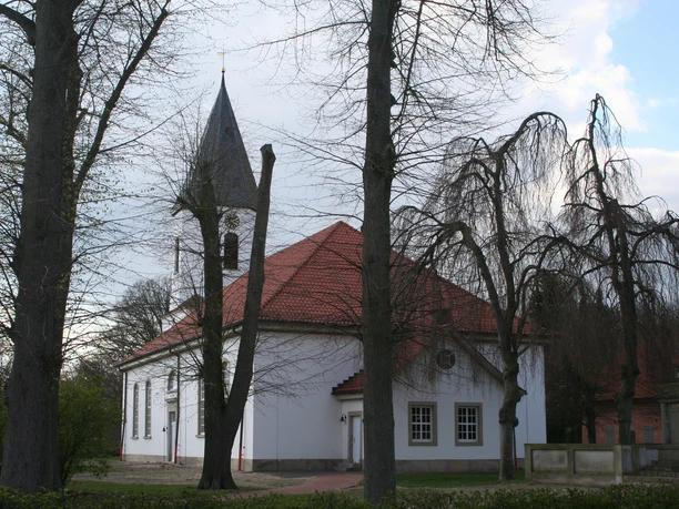 Die helle Catharinenkirche in Martfeld, umgeben von Bäumen, mit ihrem spitzen Turm und roten Dachziegeln.