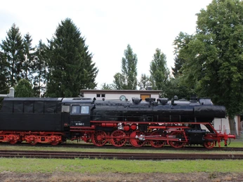 Eine historische Dampflokomotive steht auf Schienen, umgeben von Bäumen und einem Gebäude im Hintergrund.A historic steam locomotive stands on rails, surrounded by trees and a building in the background.Et historisk damplokomotiv står på skinner, omgivet af træer og en bygning i baggrunden.Een historische stoomlocomotief staat op rails, omringd door bomen en een gebouw op de achtergrond.