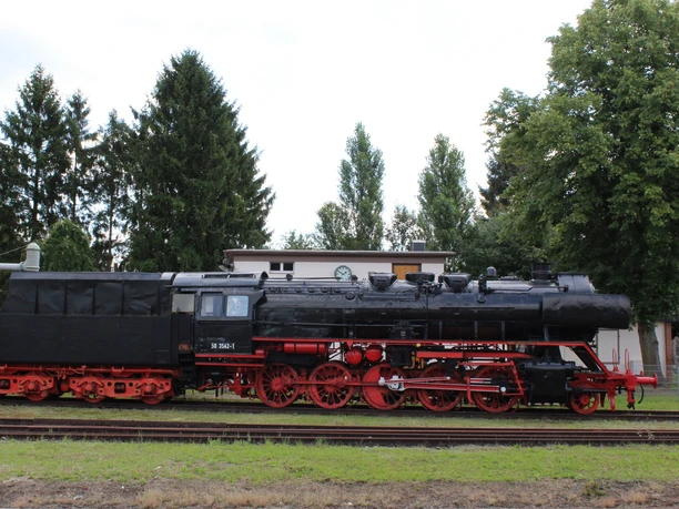 Museumslok Weyhe A historic steam locomotive stands on rails, surrounded by trees and a building in the background.