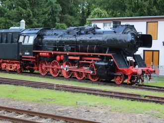 Historic steam locomotive outdoors, black with red wheels, surrounded by trees and a hut.