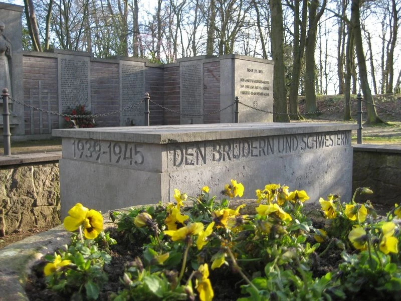 Gedenkstätte mit Blumen und Inschrift "Den Brüdern und Schwestern 1939-1945", umgeben von Bäumen.