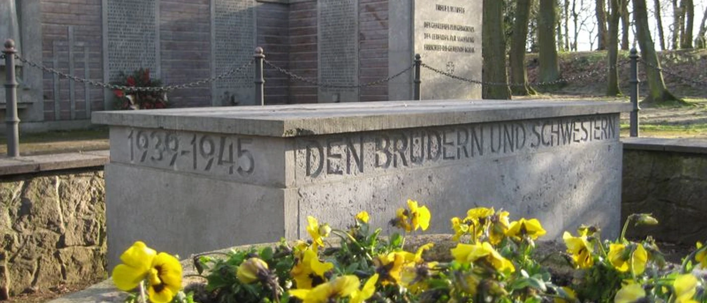 Ehrenmal Gedenkstätte mit Blumen und Inschrift "Den Brüdern und Schwestern 1939-1945", umgeben von Bäumen.