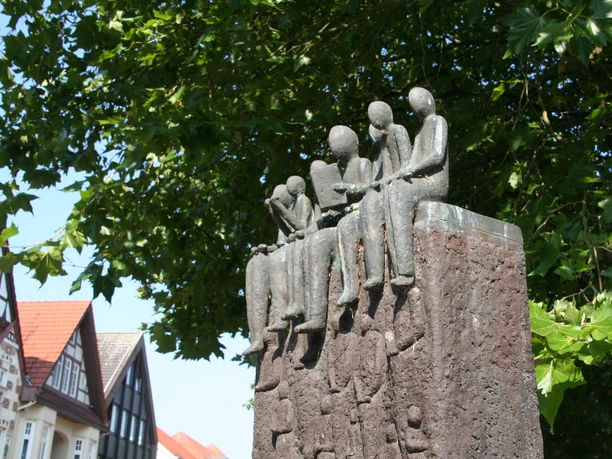 Bronzeskulptur "Der Vorleser" auf einem steinernen Sockel, umgeben von Fachwerkhäusern und Bäumen.