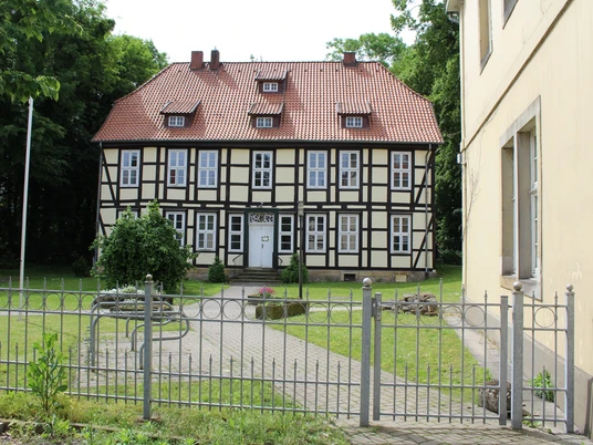 Amtshof Stolzenau Fachwerkgebäude mit rotem Ziegeldach und weißem Eingang, umgeben von einem gepflegten Garten und Zaun.
