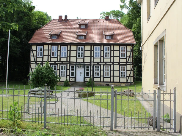 Amtshof Stolzenau Fachwerkgebäude mit rotem Ziegeldach und weißem Eingang, umgeben von einem gepflegten Garten und Zaun.