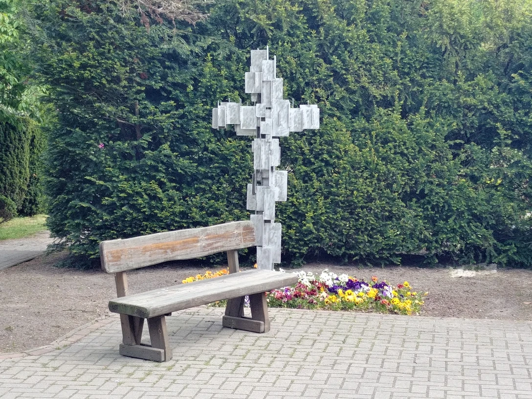 Holzbank vor Metallkreuz mit Blümchenbeet, umgeben von grünen Koniferen, ruhiger Ort.