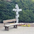 Holzbank vor Metallkreuz mit Blümchenbeet, umgeben von grünen Koniferen, ruhiger Ort.