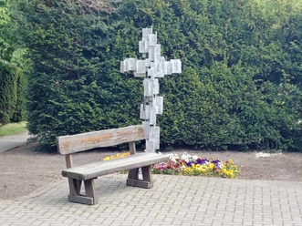 Ohne Titel Holzbank vor Metallkreuz mit Blümchenbeet, umgeben von grünen Koniferen, ruhiger Ort.