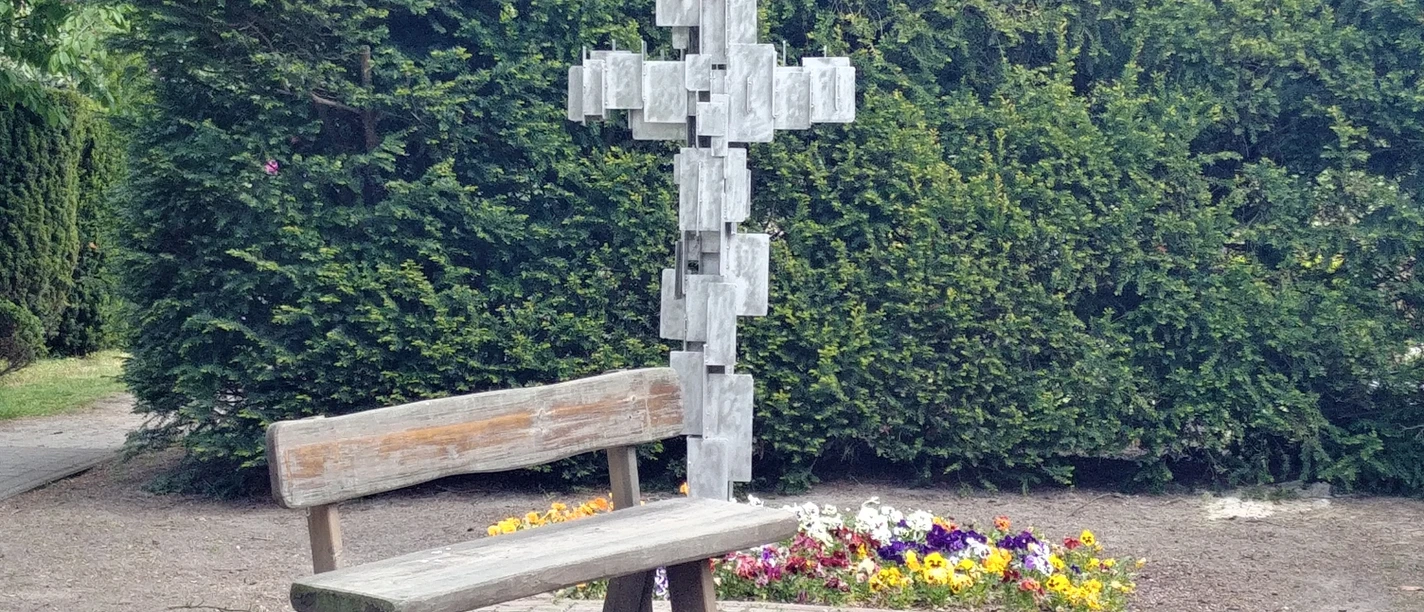 Ohne Titel Holzbank vor Metallkreuz mit Blümchenbeet, umgeben von grünen Koniferen, ruhiger Ort.