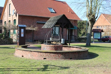Runder, gemauerter Grillplatz mit Überdachung und Holzlager, umgeben von grüner Rasenfläche.