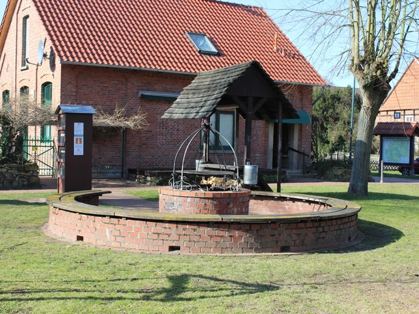 Runder, gemauerter Grillplatz mit Überdachung und Holzlager, umgeben von grüner Rasenfläche.
