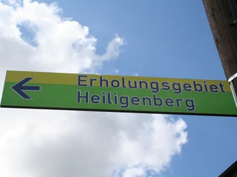 Grünes Schild mit der Aufschrift "Erholungsgebiet Heiligenberg", blauer Pfeil nach links zeigend.
